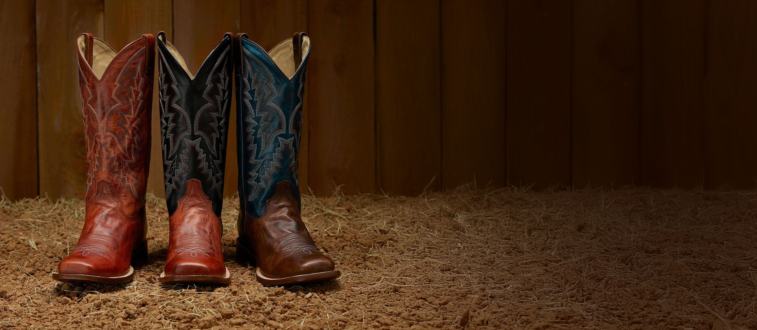 A lineup of the Camarillo 13” Western boot in brown, black and brown, and blue and brown, on a dirt floor.